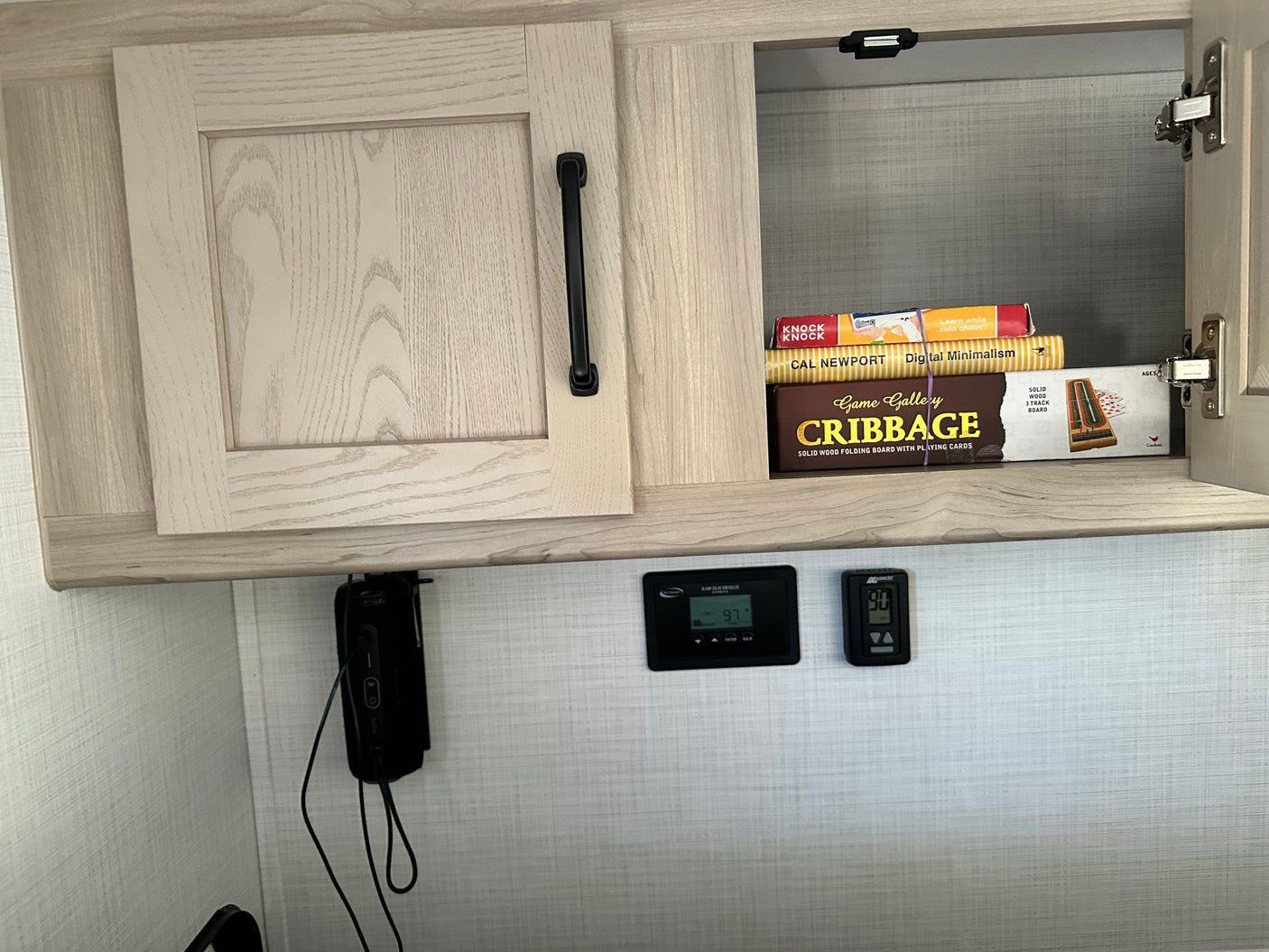 Open RV rental cabinet with games & books above a solar controller (97) and thermostat (90), showing interior storage & controls.