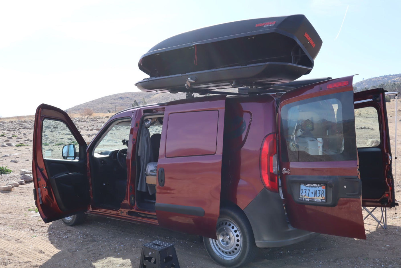 A reddish-brown camper van with all doors open and a roof cargo box, parked outdoors. Perfect for an RV rental camping trip!