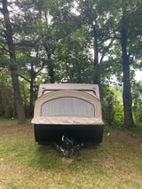 Front view of an RV rental: a beige pop-up camper parked on grass surrounded by lush green trees at a campsite.