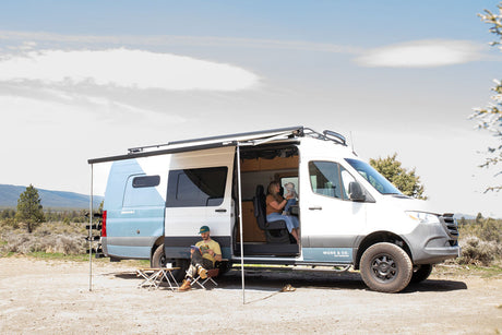 A family relaxes in an RV rental camper van in a scenic outdoor setting. A man reads outside, a woman holds a baby inside.