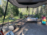 RV rental view: A serene forest campsite with picnic table, bike, and person relaxing on a blanket.