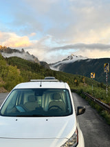 A white RV rental van parked on a scenic mountain road with misty, forested peaks and sticker-covered signs.