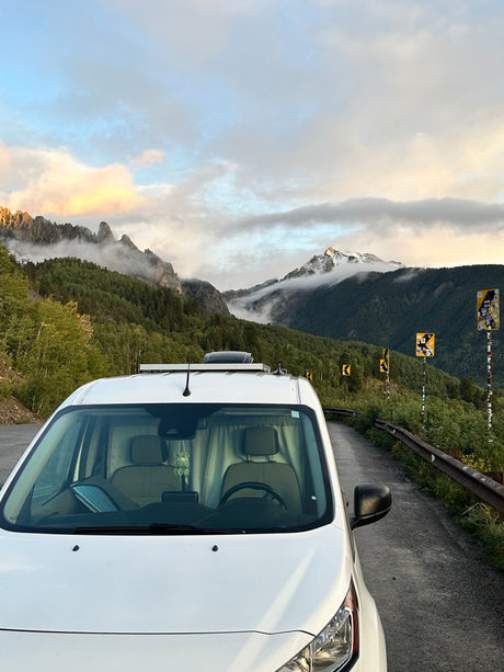 A white RV rental van parked on a scenic mountain road with misty, forested peaks and sticker-covered signs.