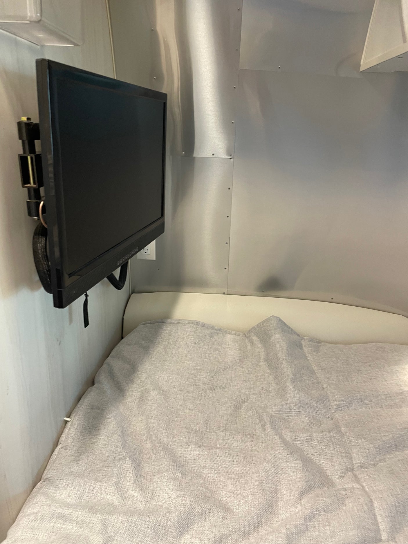 RV rental bedroom shows a wall-mounted black TV on aluminum walls, next to a bed with a light grey blanket.