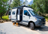 Winnebago RV rental camper van parked in nature. Side door open, revealing interior, with outdoor table, drinks & stools.