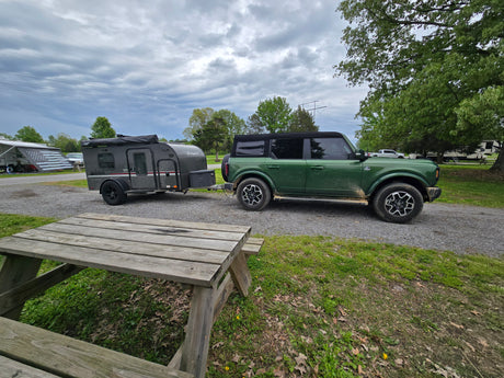Green Bronco and a tear-drop RV rental trailer parked at a gravel campsite with a picnic table. Perfect for camping adventures.