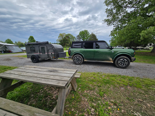 Green Bronco and a tear-drop RV rental trailer parked at a gravel campsite with a picnic table. Perfect for camping adventures.