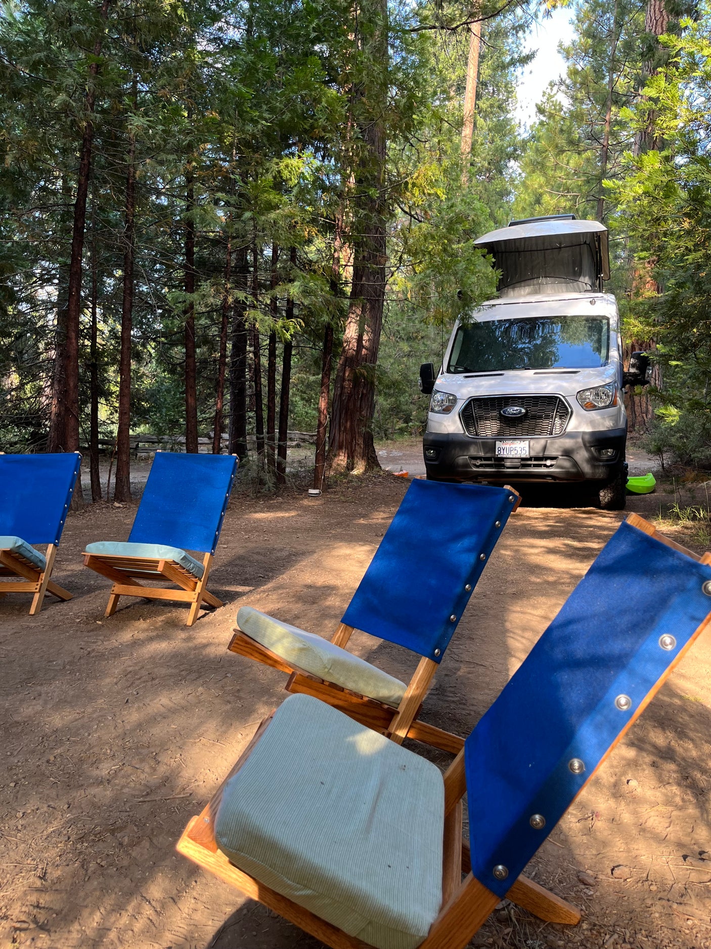 White RV rental van at a forest campsite. Blue folding chairs with light green cushions invite relaxation in nature.
