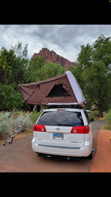 Ready for an rv rental adventure? See this white minivan with a brown rooftop tent & ladder, setup for camping by a red rock mountain.