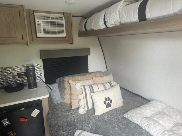 Cozy RV rental interior with a dog-friendly sleeping area, kitchenette, AC unit, and overhead bunk bed. Compact & inviting.