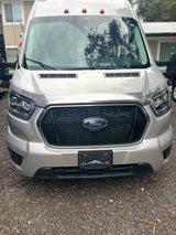 Silver Ford Transit van, likely an RV rental, shown from the front with a unique mountain-themed license plate frame.