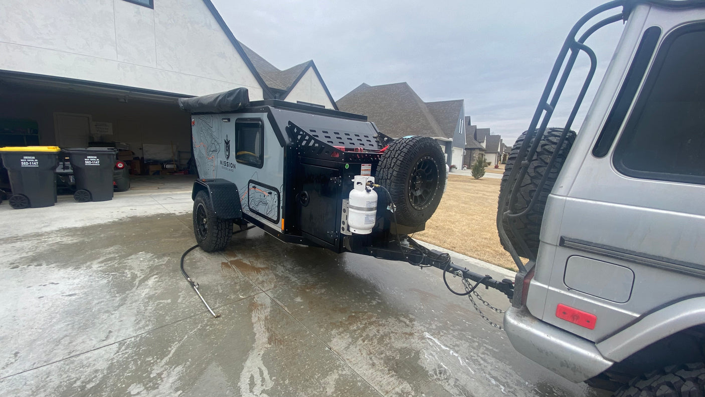 An overland adventure trailer, an ideal RV rental, is hitched to an SUV on a wet driveway, ready for travel.