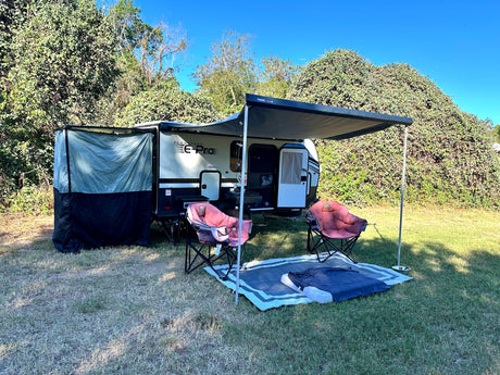 An RV rental, a Flagstaff E-Pro, is set up on a grassy site with its awning, side tent, chairs, and a mat under blue sky.
