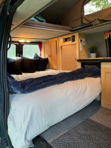 RV rental interior: large bed with blue blanket, wood cabinets, microwave, and pop-top roof for extra space.