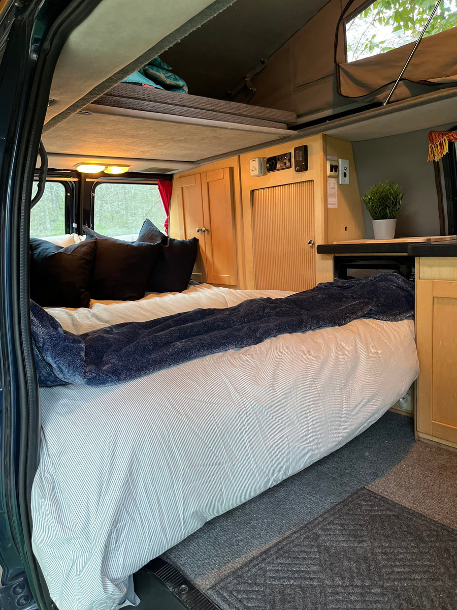 RV rental interior: large bed with blue blanket, wood cabinets, microwave, and pop-top roof for extra space.