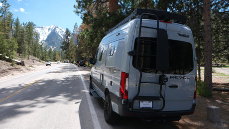 Adventure awaits! Grey Winnebago Revel rv rental parked by a mountain road, with snowy peaks & forest. Ideal for nature exploration.