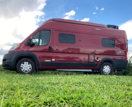 Side view of a red Winnebago Solis camper van, ideal for RV rental, parked on vibrant green grass under a partly cloudy sky.
