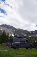 Dark gray RV rental van with bikes, parked in a mountain meadow with yellow flowers & snow-capped mountains.