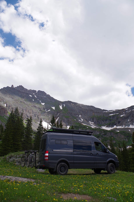 Dark gray RV rental van with bikes, parked in a mountain meadow with yellow flowers & snow-capped mountains.