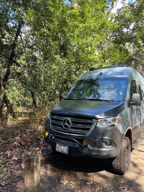 Adventure-ready Mercedes Sprinter van, perfect for an RV rental, parked on a dirt path in a sunny forest.