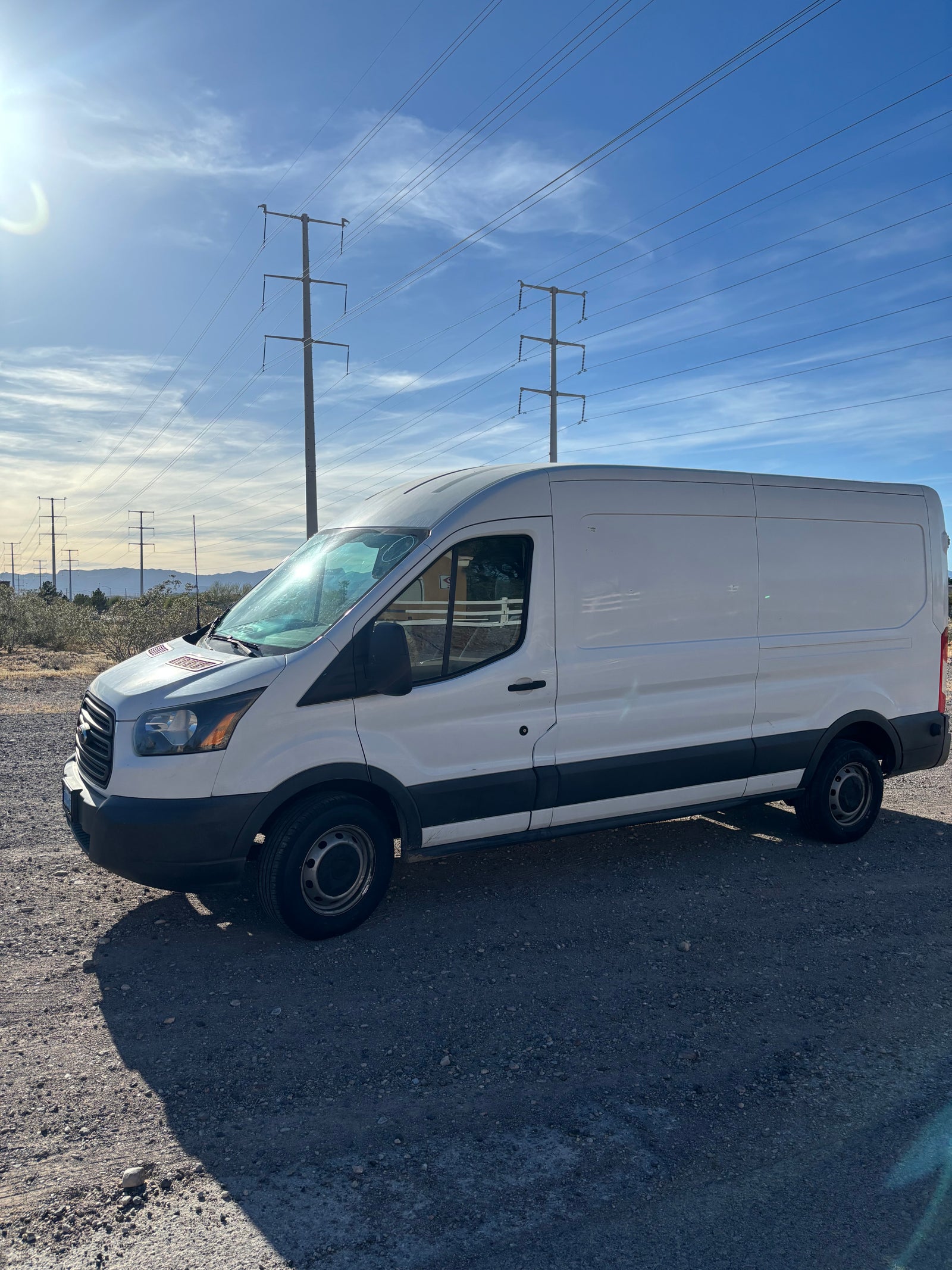 White cargo van, potentially an RV rental, parked on gravel under power lines with mountains in background.