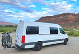 Scenic RV rental with two bikes, parked at a trailhead against a backdrop of red rock mountains and a green valley.