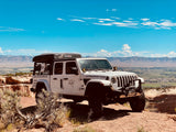 This white Jeep Gladiator **RV rental** with a rooftop tent overlooks a scenic valley from a desert cliff, ready for adventure.