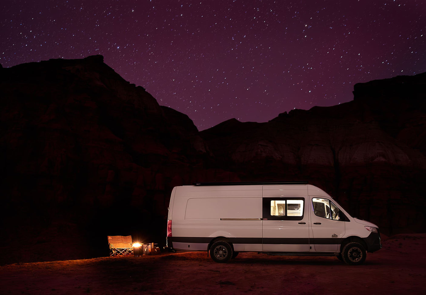 RV rental van under star-dusted purple night sky. Illuminated camper with a cozy campfire & chairs in a dark landscape.