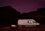 RV rental van under star-dusted purple night sky. Illuminated camper with a cozy campfire & chairs in a dark landscape.