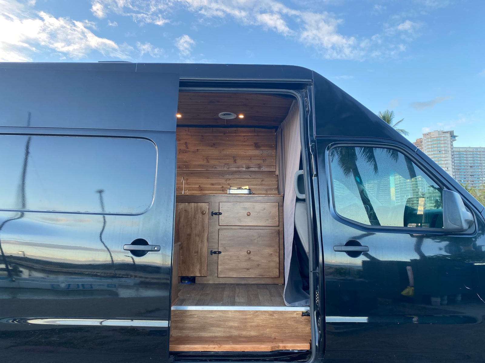 Explore this custom black camper van, perfect as an rv rental. Its open side reveals a cozy wooden kitchen & storage area.