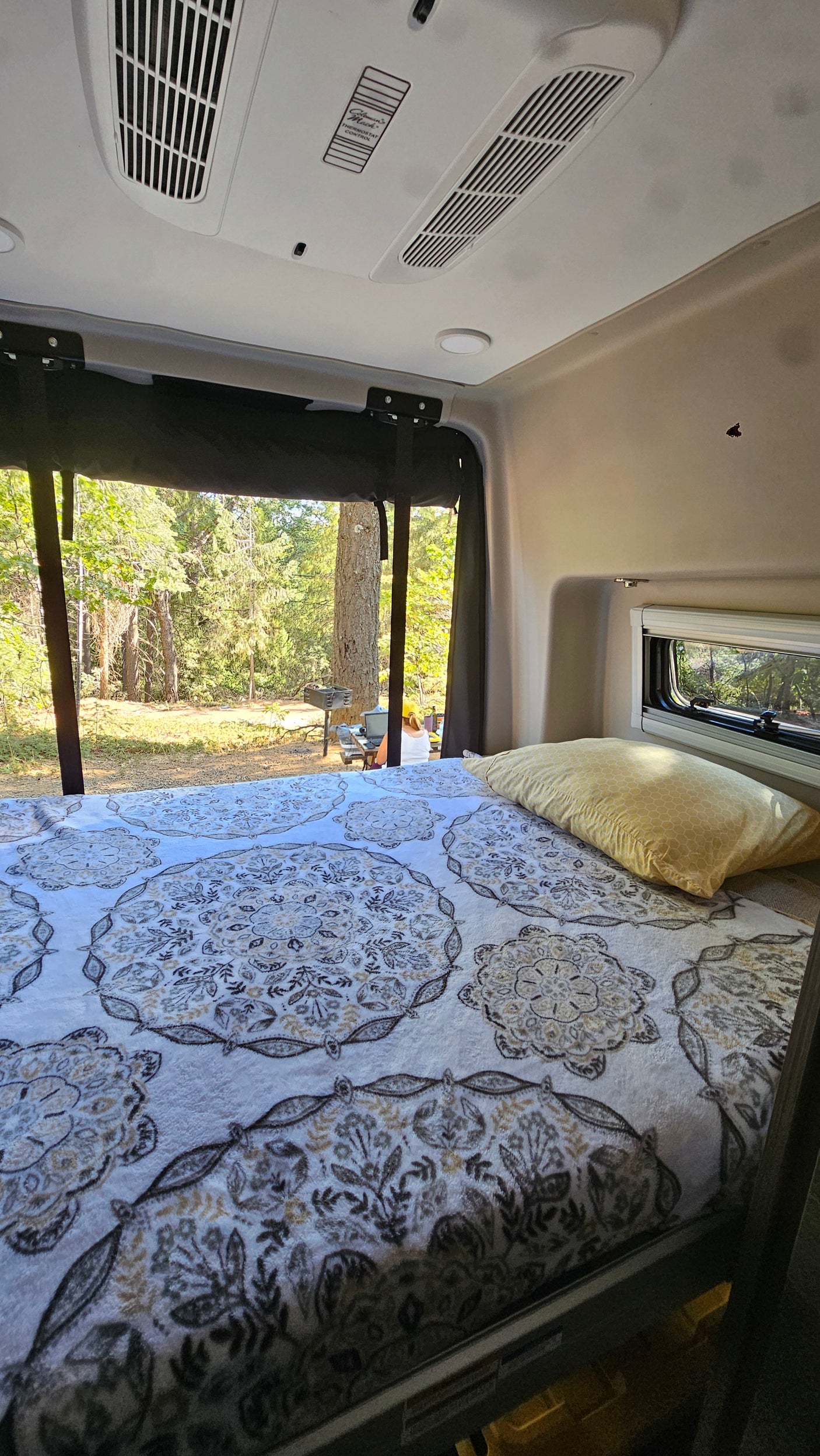 Cozy RV rental bedroom with a large window revealing a serene forest view. Features a patterned blanket, yellow pillow, and overhead AC.
