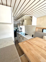 Bright RV rental interior with a sleek kitchenette, wood-top table, and comfy bench seating. Ideal for mobile living.