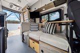 Cozy RV rental interior view from the rear, showing comfortable seating, a mounted TV, and a scenic lake and beach.