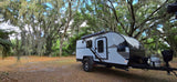 A white Bushwhacker travel trailer, available for RV rental, is parked under magnificent moss-covered trees.