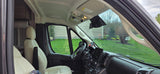 Interior view of an RV rental cockpit featuring light-colored seats, a modern dashboard, and a yard view through the windshield.