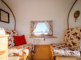 Cozy wagon-style RV rental interior features patterned benches, a fold-down table, window with curtains, and rustic decor.