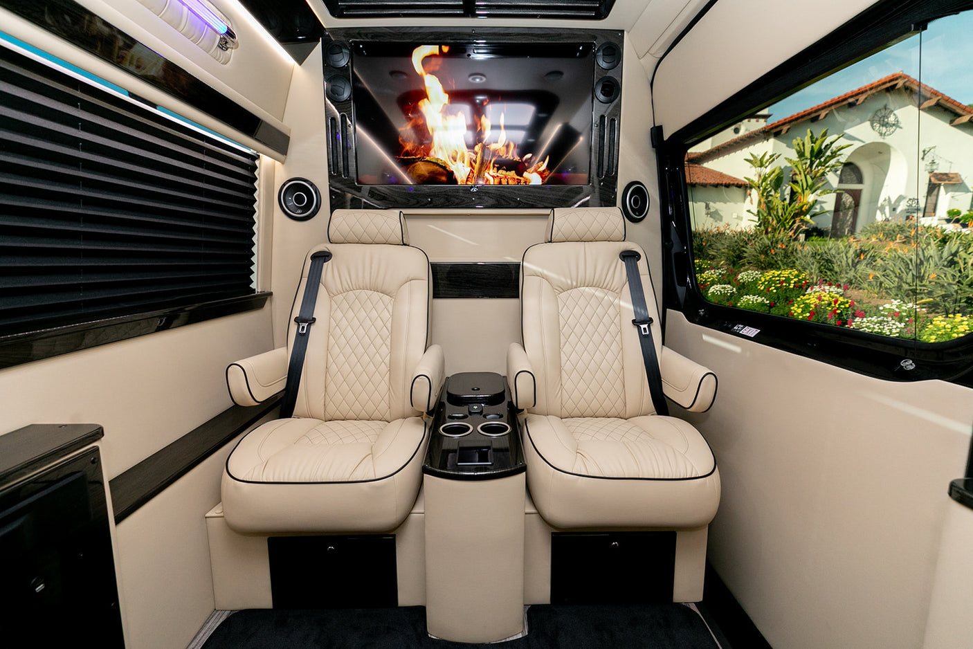 Luxury RV rental interior featuring cream quilted leather captain's chairs, center console, large screen with a digital fire, and window views.