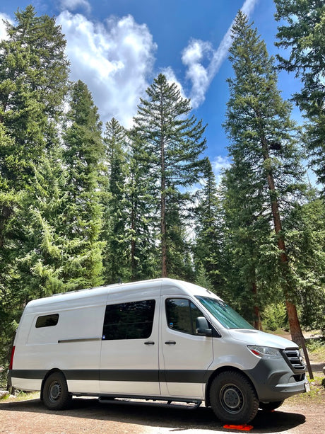 A white Sprinter camper van is parked among tall pine trees under a blue sky. Perfect RV rental for nature adventures.