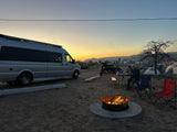 Campsite at sunset: an RV rental, warm campfire, and people enjoying the desert evening amidst tents and vehicles.