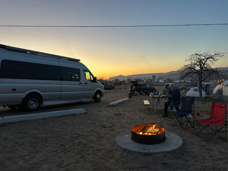 Campsite at sunset: an RV rental, warm campfire, and people enjoying the desert evening amidst tents and vehicles.