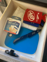 A well-stocked drawer in an RV rental with a first aid kit, cutting board, knife, and drink koozies for travel essentials.