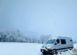 Experience winter magic! A grey rv rental van is parked in a deep snowfield, surrounded by frost-covered trees under a soft, snowy sky.