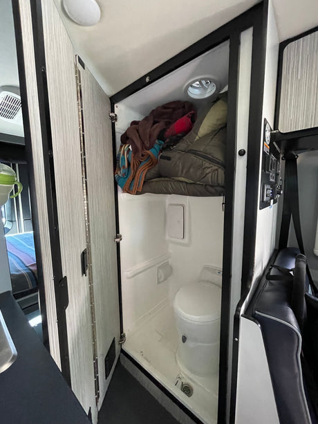 An RV rental's compact bathroom is revealed with a toilet, shower drain, and practical overhead storage.