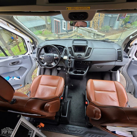 Inside a modern RV rental van: spacious front cabin with brown leather captain's chairs and a clean dashboard.
