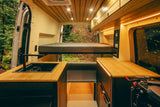 Inside a custom RV rental, this spacious camper van features a raised bed, kitchen, and forest views from the open rear.