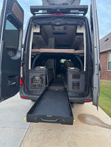 Rear view of an RV rental camper van with open doors, extended storage tray, utility access, and interior bed setup visible.