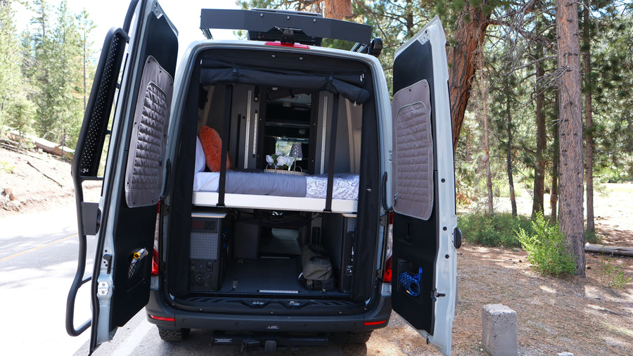 Back of an rv rental camper van open in a forest, revealing a custom bed, storage, and adventure-ready interior.