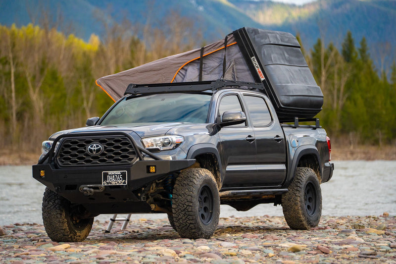 Gray Toyota Tacoma pickup with a Roofnest rooftop tent by a river. An ideal RV rental for rugged overlanding adventures.|A dark gray Tacoma truck with an open rooftop tent sits on rocky ground — photo 1