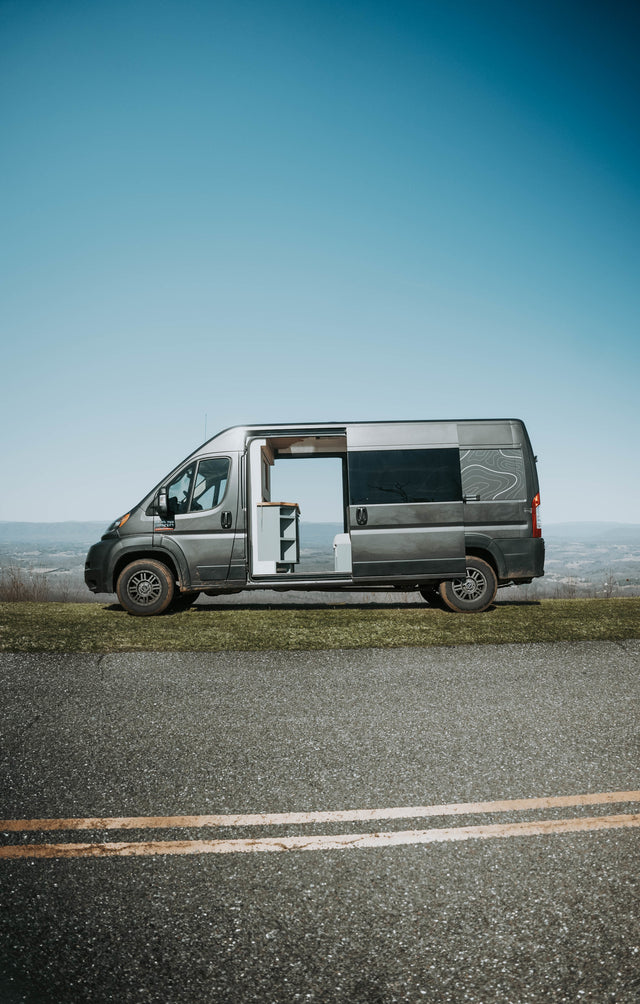 Grey camper van (RV rental) on a scenic overlook with an open side door, ready for adventure.
