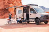 RV rental adventure: A couple enjoys outdoor cooking and relaxation next to their beige camper van in a stunning red rock desert.